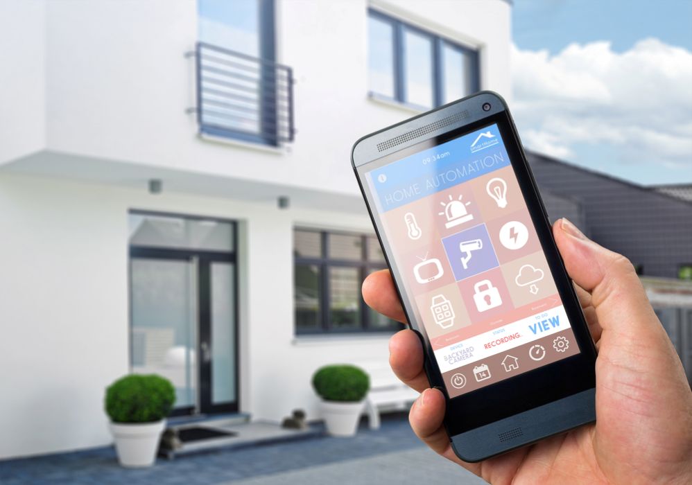 A person outside a modern house holding a smartphone showing a home automation app to control smart alarm and security features.