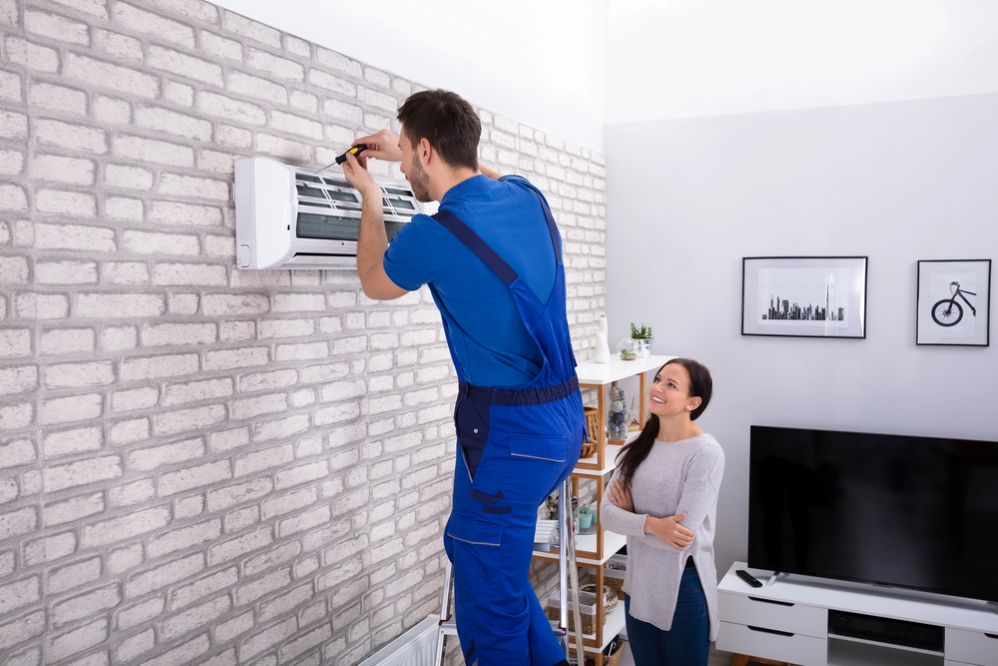 A technician installing a wall-mounted heat pump while a homeowner looks on in a modern living room.