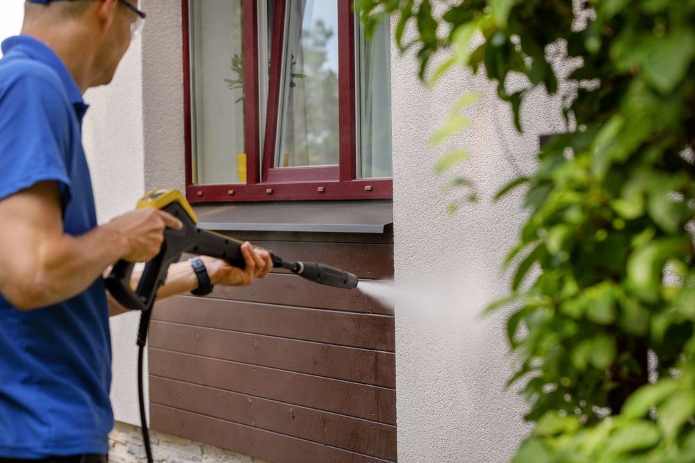 A person pressure washing exterior weatherboard near a window on a residential home.