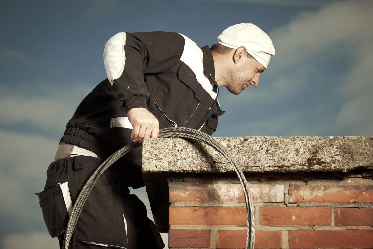 A professional chimney sweep inspecting and cleaning a brick chimney using a steel brush cable.