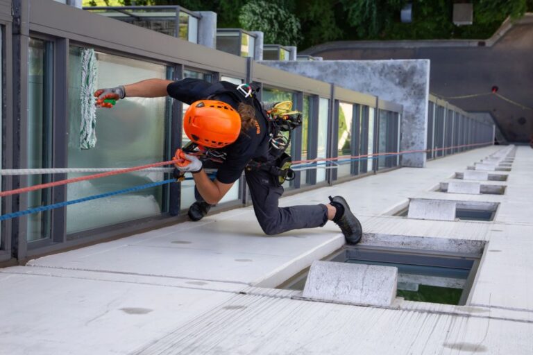 A rope access technician in full safety gear cleaning high-rise windows, securely suspended by dual ropes and working across the building façade.