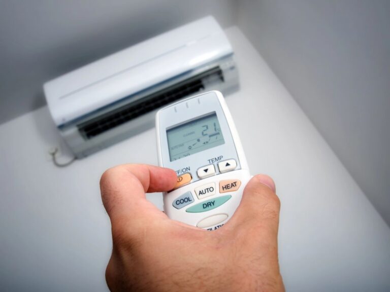 A hand holding a heat pump remote set to 21 degrees while operating a wall-mounted heat pump unit.