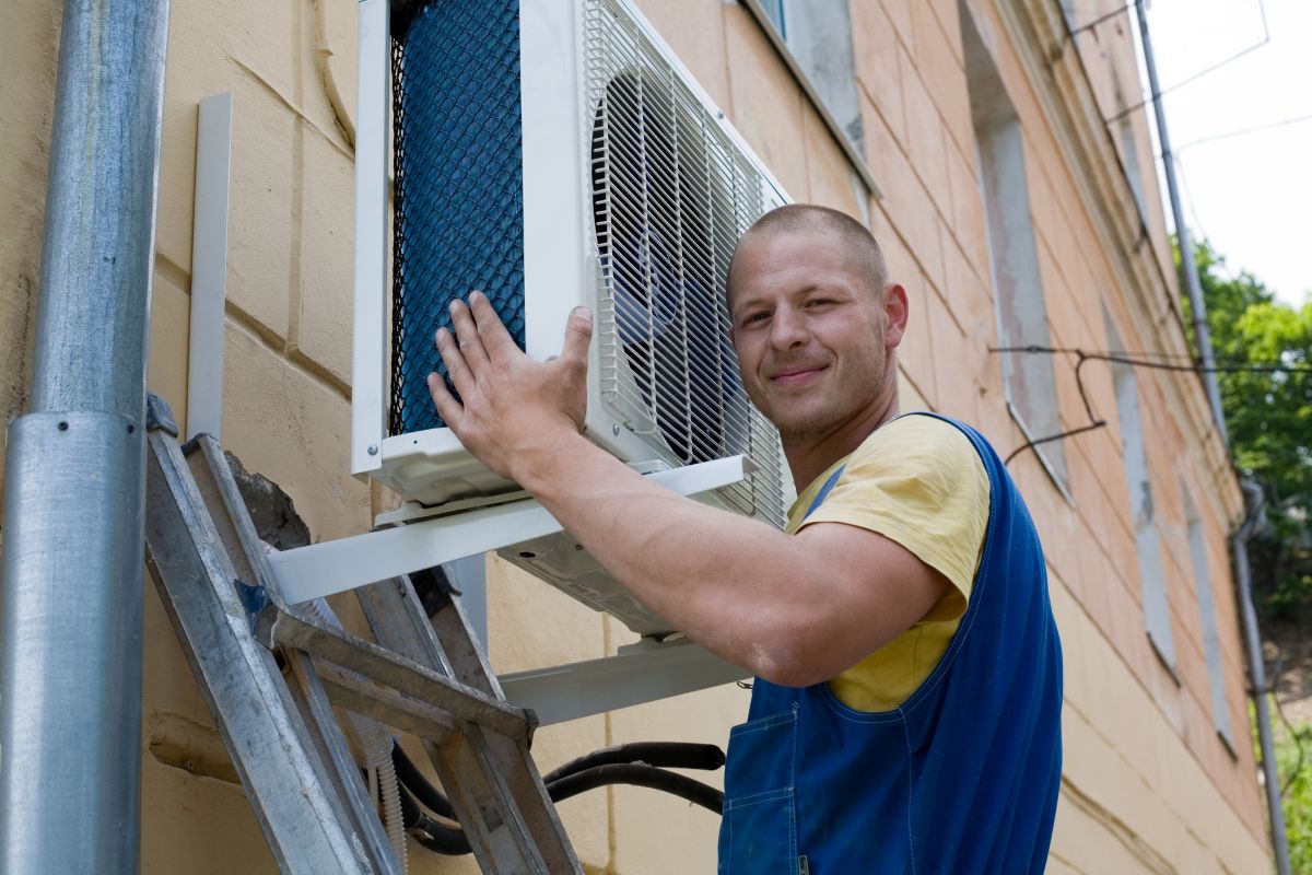 A technician installing an outdoor air conditioning unit on a wall bracket outside a home.